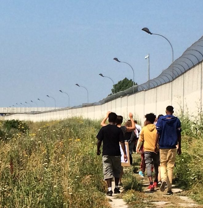groupe d'ados marchant sur un chemin avec à gauche des herbes et fleurs, à droite un mur surmonté de barbelé, à Calais